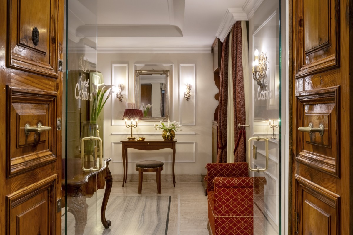 Vista prospettica dell'ingresso dell'Hotel Cortina a Roma, attraverso porte di vetro che riflettono la hall e portano a un'area salotto - Hotel Cortina, BZAR hotels a Roma