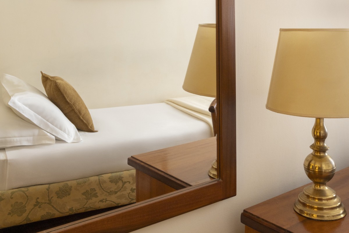 Dettaglio di una camera Small accogliente e ben organizzata all'Hotel Cortina a Roma, con letto singolo, scrivania in legno antico, sedia decorata e specchio che riflette la luce della finestra - Hotel Cortina, BZAR hotels a Roma