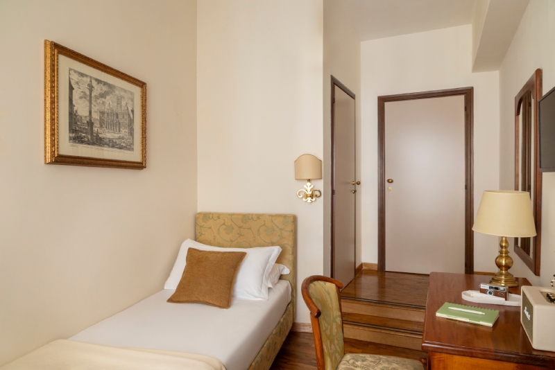 Bright single room at Hotel Cortina in Rome, featuring classic decor with a wooden desk, vintage chair, suitcase by the window, and a single bed with crisp white linens - Hotel Cortina, BZAR hotels a Roma