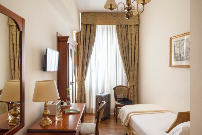Una luminosa camera singola dell'Hotel Cortina a Roma, arredata in stile classico con scrivania in legno, sedia d'epoca, valigia accanto alla finestra e letto singolo con biancheria bianca - Hotel Cortina, BZAR hotels a Roma