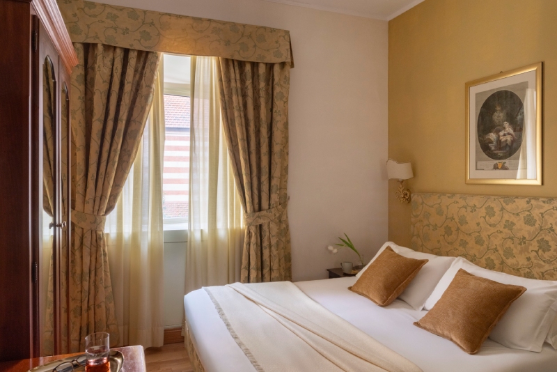 Vista serena di una camera Small presso l'Hotel Cortina a Roma, con un letto singolo dal design elegante, cuscini in velluto marrone, un armadio d'epoca e una grande finestra con tende fiorite e trasparenti - Hotel Cortina, BZAR hotels a Roma