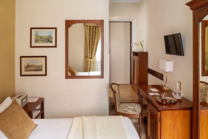Dettaglio di una camera Small accogliente e ben organizzata all'Hotel Cortina a Roma, con letto singolo, scrivania in legno antico, sedia decorata e specchio che riflette la luce della finestra - Hotel Cortina, BZAR hotels a Roma