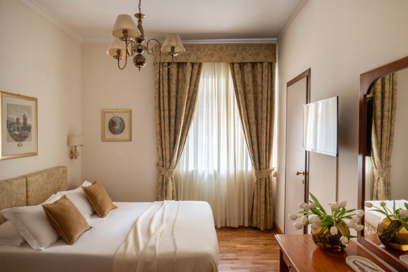 Accogliente camera standard dell'Hotel Cortina a Roma, caratterizzata da un comodo letto matrimoniale con cuscini marroni, pavimento in parquet, e una grande finestra con eleganti tende fiorite - Hotel Cortina, BZAR hotels a Roma
