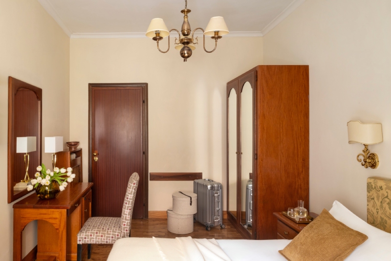 Interno spazioso di una camera standard presso l'Hotel Cortina a Roma, con una scrivania in legno d'epoca, una sedia imbottita, un grande armadio a muro e una valigia trolley silver - Hotel Cortina, BZAR hotels a Roma